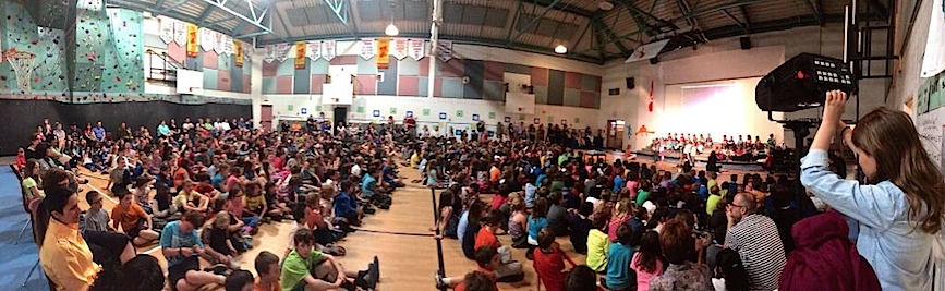 The school is assembled in the gym. A girl holds a spotlight pointing to people seated on benches in front of the crowd.