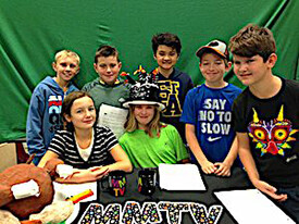 Seven kids sit a table with some mugs and papers. There's a green screen behind them. Letters on the table read "MMTV."